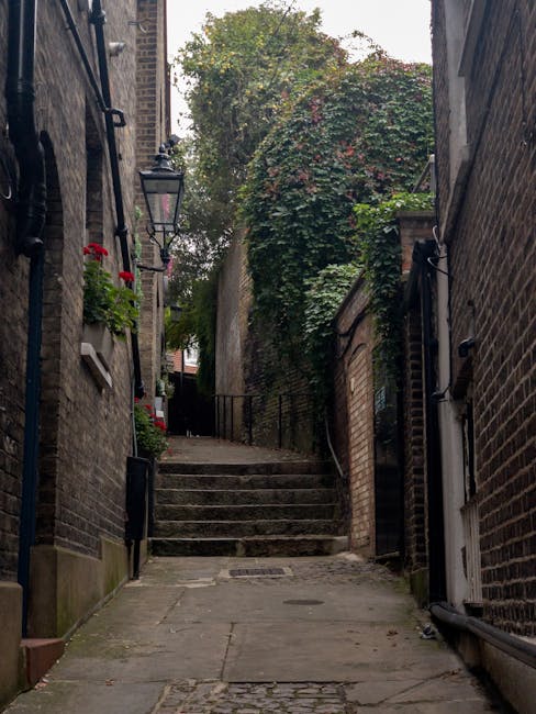 A narrow cobblestone alleyway between historic brick buildings in Golders Green, featuring a slight incline with stone steps at the top. On one side, a black wrought-iron lantern is mounted on the bri