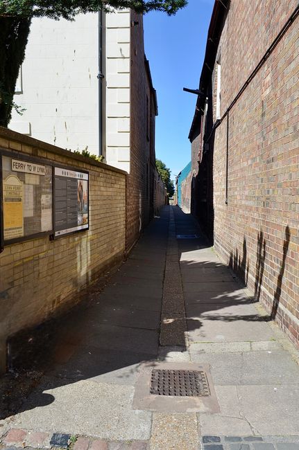 A narrow cobblestone alleyway between historic brick buildings in Golders Green, featuring a slight incline with stone steps at the top. On one side, a black wrought-iron lantern is mounted on the bri